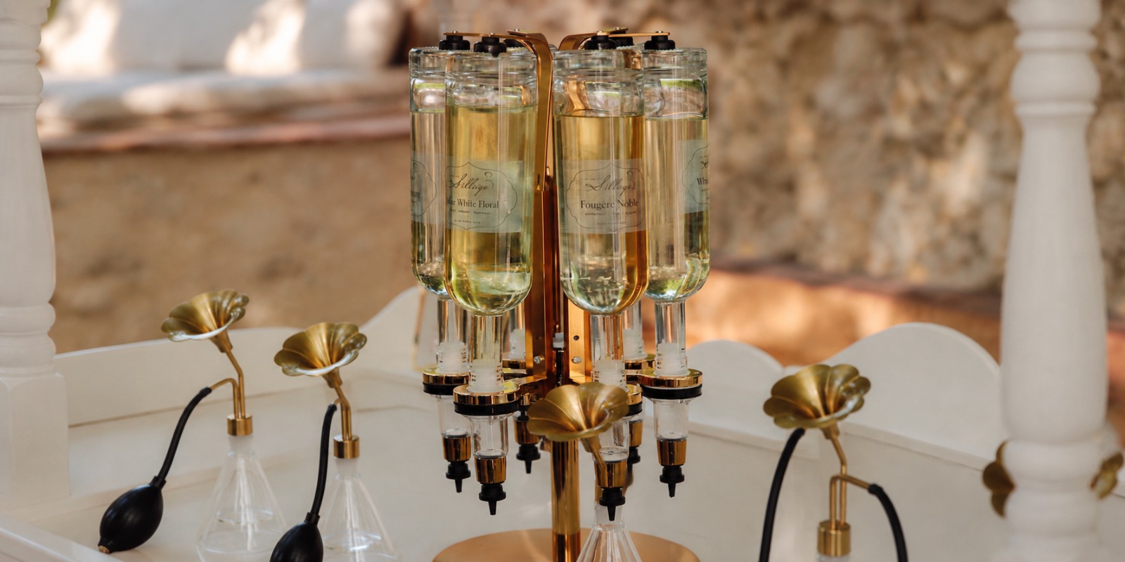 Carrousel doré de flacons de parfum et vaporisateurs à poire noire sur une table blanche, devant un mur en pierre ensoleillé.