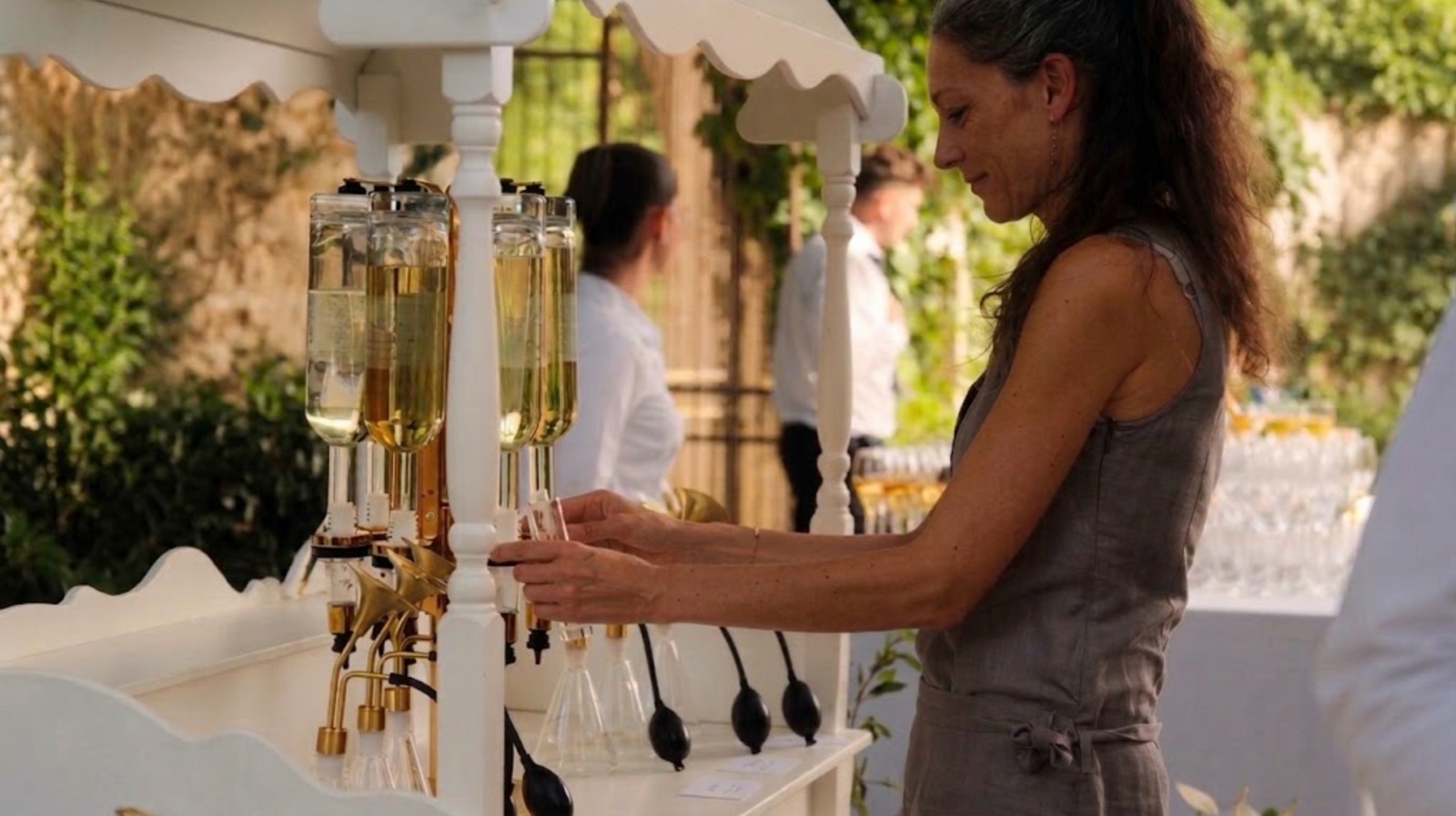 La parfumeuse sert une fragrance depuis le bar à parfums blanc et laiton, dans le jardin d'une réception de mariage.