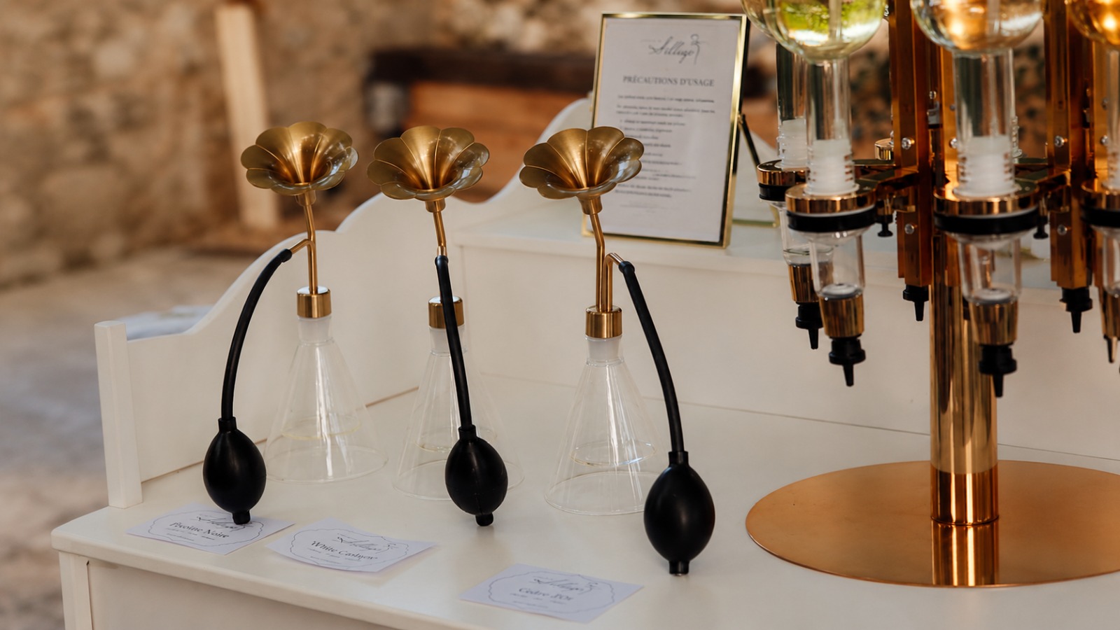 Vaporisateurs vintage à têtes dorées, entonnoirs en verre et cartes parfumées disposés sur une table blanche.